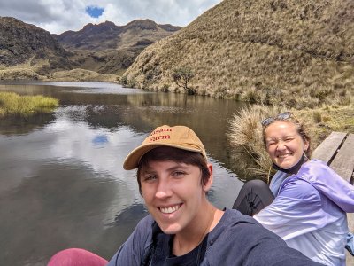 Two young woman smile at the camera. In the background is a river and mountains.