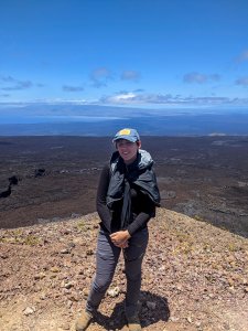 A woman in a baseball cap, black shirt and gray pants stands on a mountaintop.